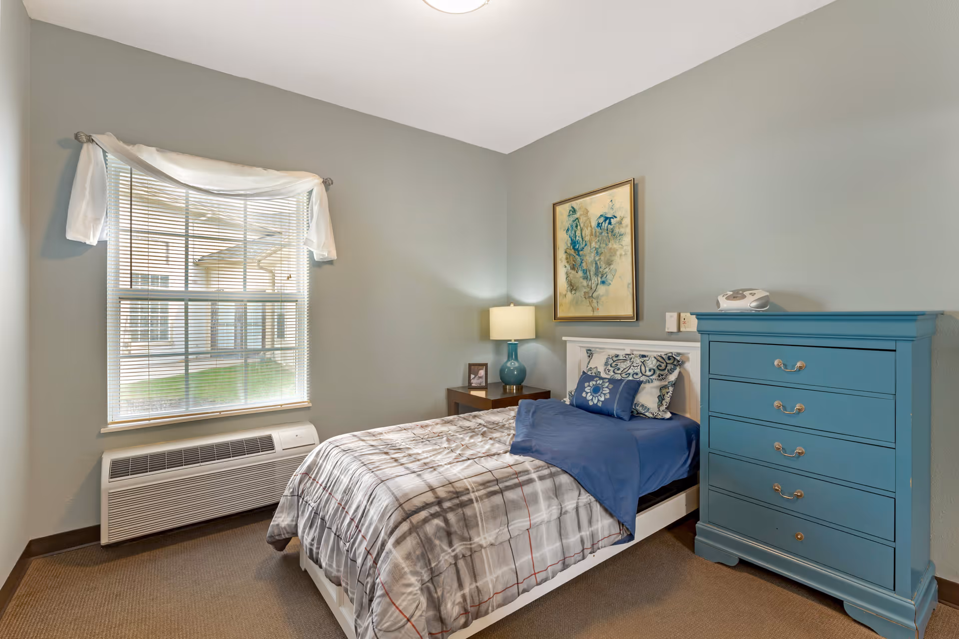 A neatly made single bed with a plaid comforter and blue pillows in a senior living bedroom. Next to the bed is a wooden nightstand with a blue lamp and a framed photo. A blue chest of drawers stands against the wall. A window with white curtains lets in natural light, and a framed abstract painting hangs above the bed.