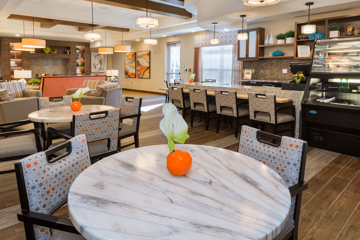 A bright and modern dining area in a senior living facility featuring round marble tables with patterned chairs, a long counter with bar stools, pendant lighting, and a display case with beverages and snacks. The room has warm wood flooring, decorative wall art, and a cozy seating area with sofas and lamps in the background.