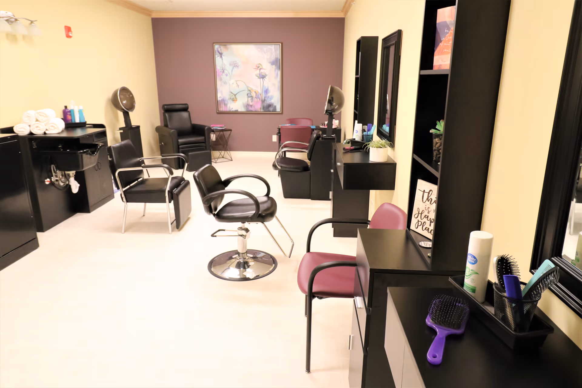 Interior view of a salon area in a senior living facility with black salon chairs, hair washing station, mirrors, and shelves with hair care products and brushes. The walls are painted beige and purple, and there is a framed floral artwork on the purple wall.
