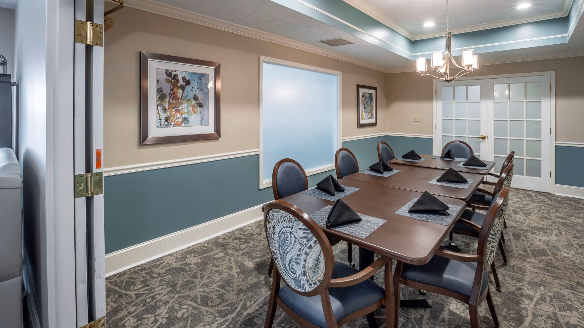 A dining room with a rectangular wooden table set for eight people. Each place setting has a gray placemat and a black folded napkin. The chairs have wooden frames with blue upholstered seats and backs, some with patterned fabric on the backrest. The room has beige walls with a blue wainscoting and white trim. Two framed abstract paintings hang on the walls, and a chandelier with five lights hangs above the table. Double glass-paneled doors are visible at the far end of the room.
