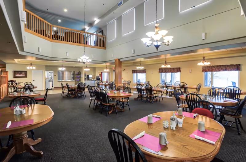 Spacious dining room with multiple round wooden tables set with pink placemats, napkins, cups, and condiments. The room has high ceilings with hanging chandeliers, large windows with valances, and a second-floor balcony overlooking the area.