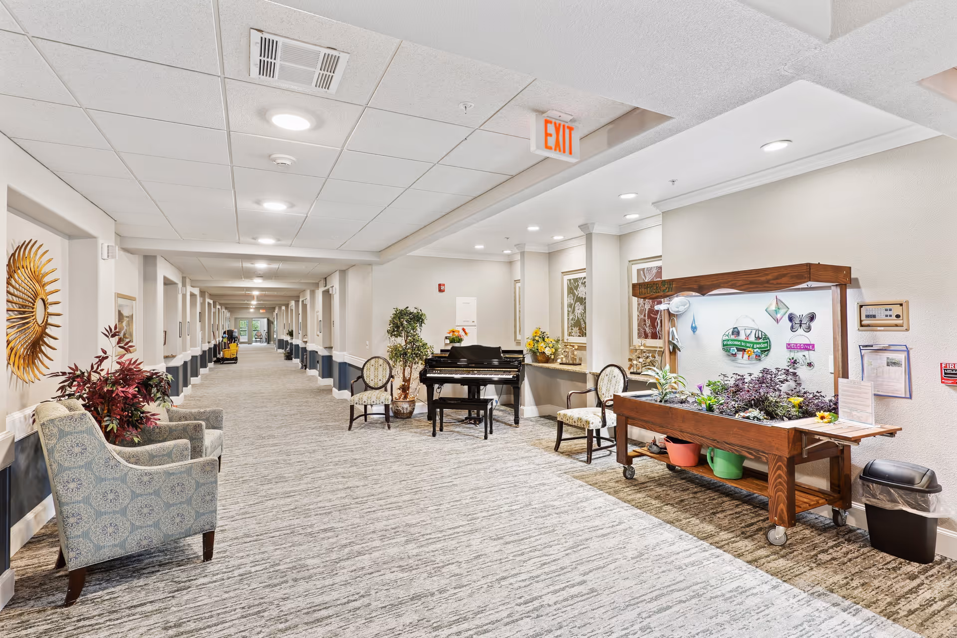 Bright, carpeted senior living facility hallway with seating, a piano, and a plant display table under an EXIT sign.