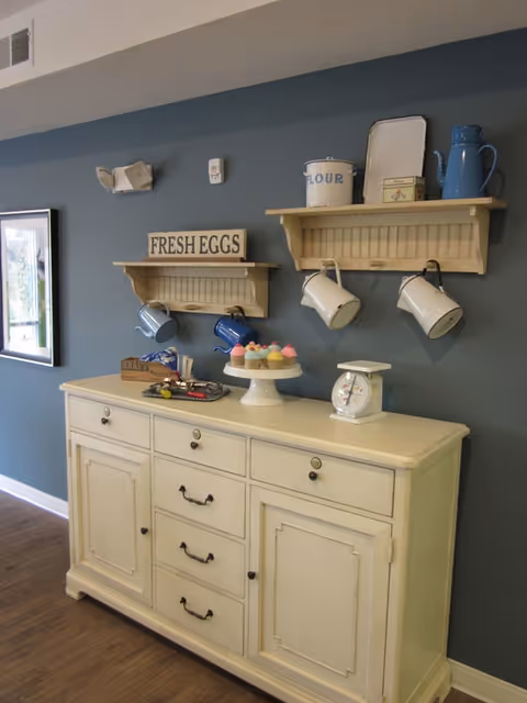 A cream-colored sideboard with drawers and cabinets against a blue wall. Above it are two wooden shelves holding decorative items including mugs, a container labeled 'FLOUR', a blue pitcher, and a sign that says 'FRESH EGGS'. On the sideboard are a white kitchen scale and a cake stand with colorful cupcakes. A framed picture is partially visible on the left wall.