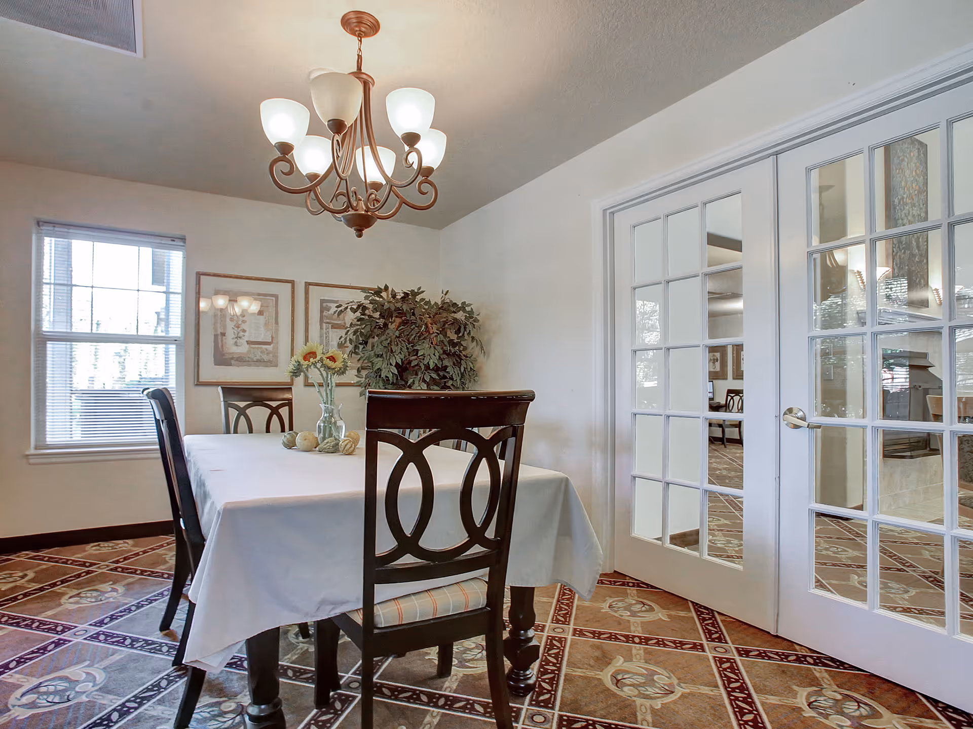 Dining room with a white-clothed table, dark wooden chairs, a chandelier, framed artwork, potted plant, and glass French doors.
