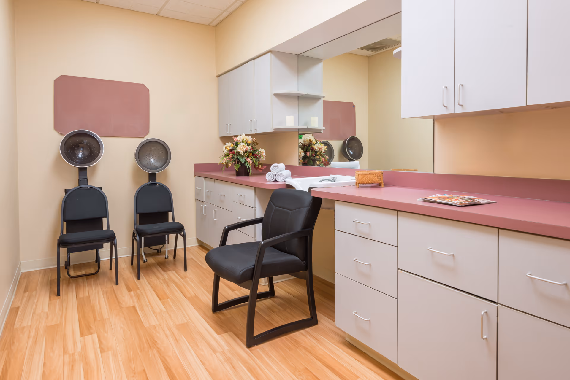 Interior view of a salon area in a healthcare facility featuring two black hair dryer chairs against a beige wall, a black armchair in front of a long counter with pink countertops, white cabinets, a large mirror, a vase with flowers, and neatly rolled white towels.