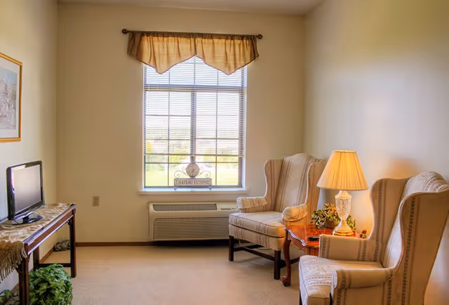 A cozy living room with two beige wingback chairs, a wooden side table with a lamp and a small plant, a window with blinds and a valance, a small TV on a wooden table with a lace runner, and a framed picture on the wall.