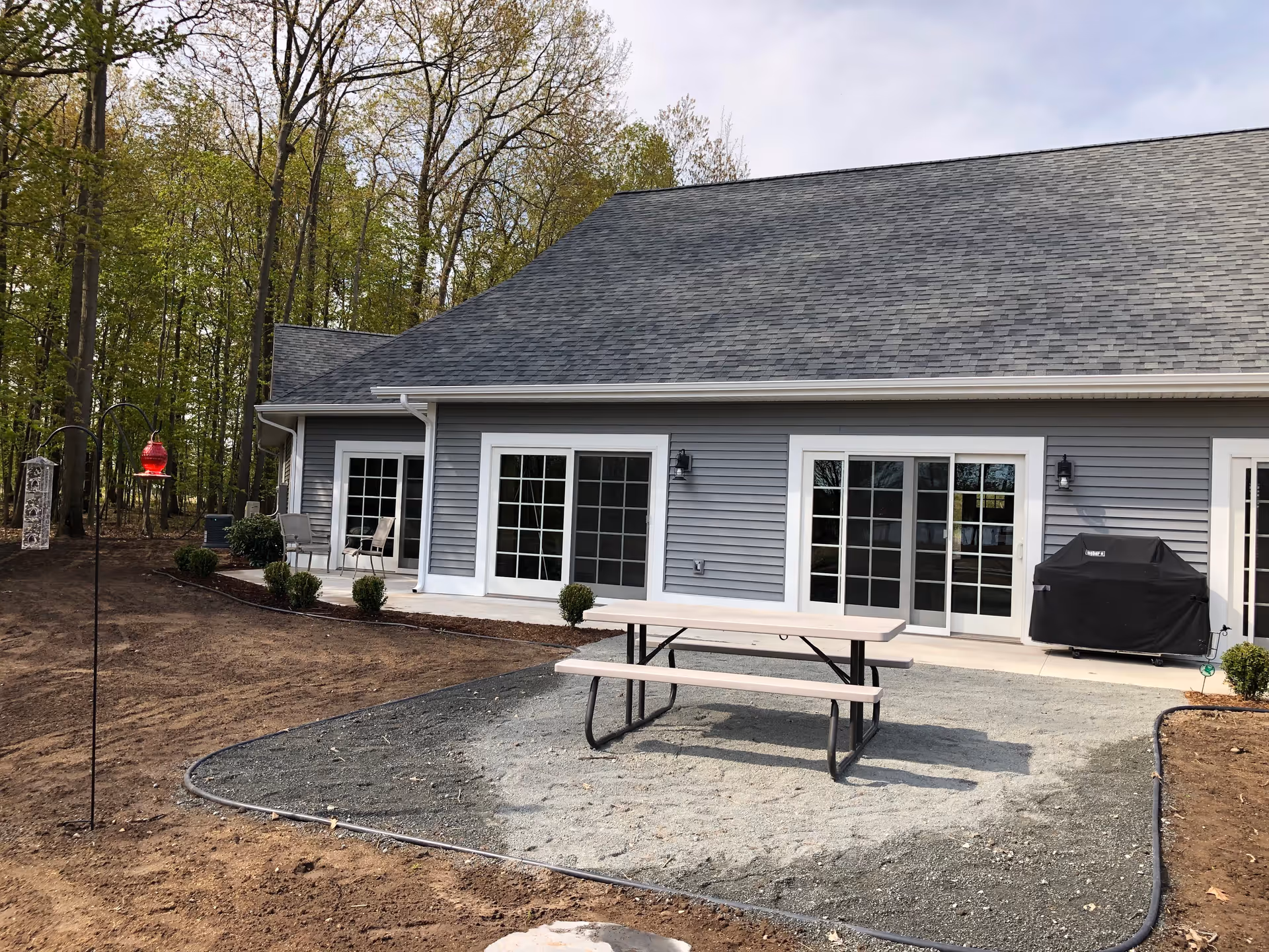 Outdoor patio area at The Cottages At Martin Lake featuring a picnic table on a gravel surface, a covered grill, and a building with multiple large windows and sliding glass doors. The area is bordered by small bushes and surrounded by trees.