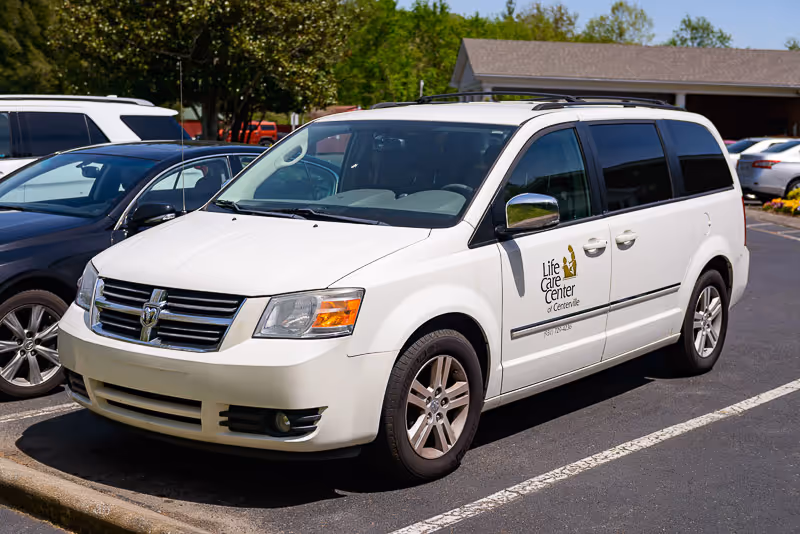White Dodge minivan parked in a parking lot with the logo and text 'Life Care Center of Centerville' on the front passenger door. Other cars and trees are visible in the background.