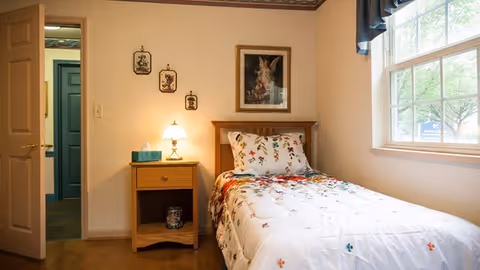 A small bedroom with a single bed covered in a white floral comforter and matching pillow. Next to the bed is a wooden nightstand with a lamp and a box of tissues. On the wall above the bed are three small framed pictures and one larger framed picture. A window with a dark valance lets in natural light, and an open door reveals a glimpse of another room.