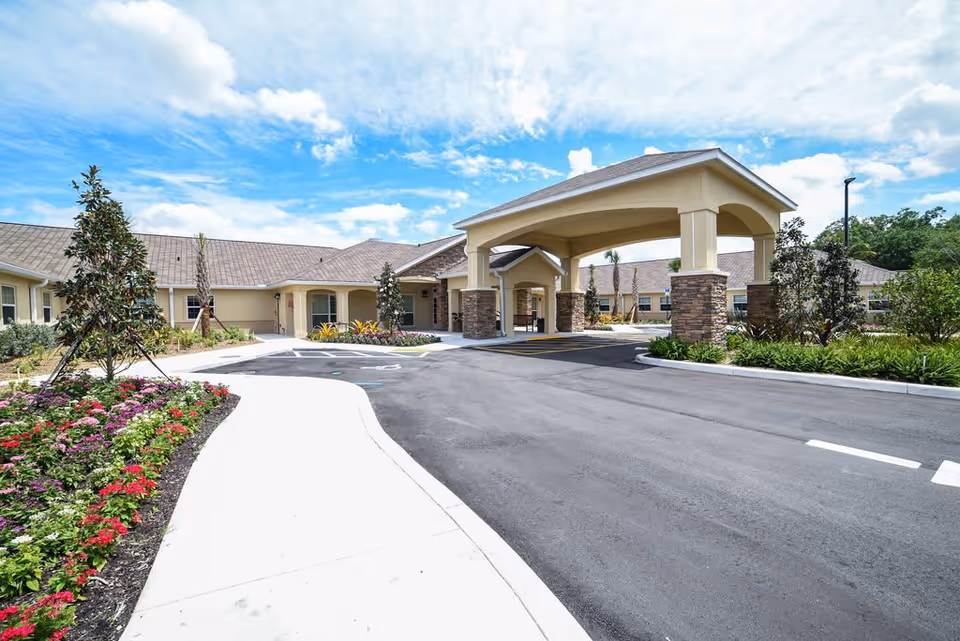 Exterior view of Artis Senior Living of Davie showing a driveway leading to a covered entrance with stone pillars, surrounded by landscaped gardens with flowers and trees under a partly cloudy sky.