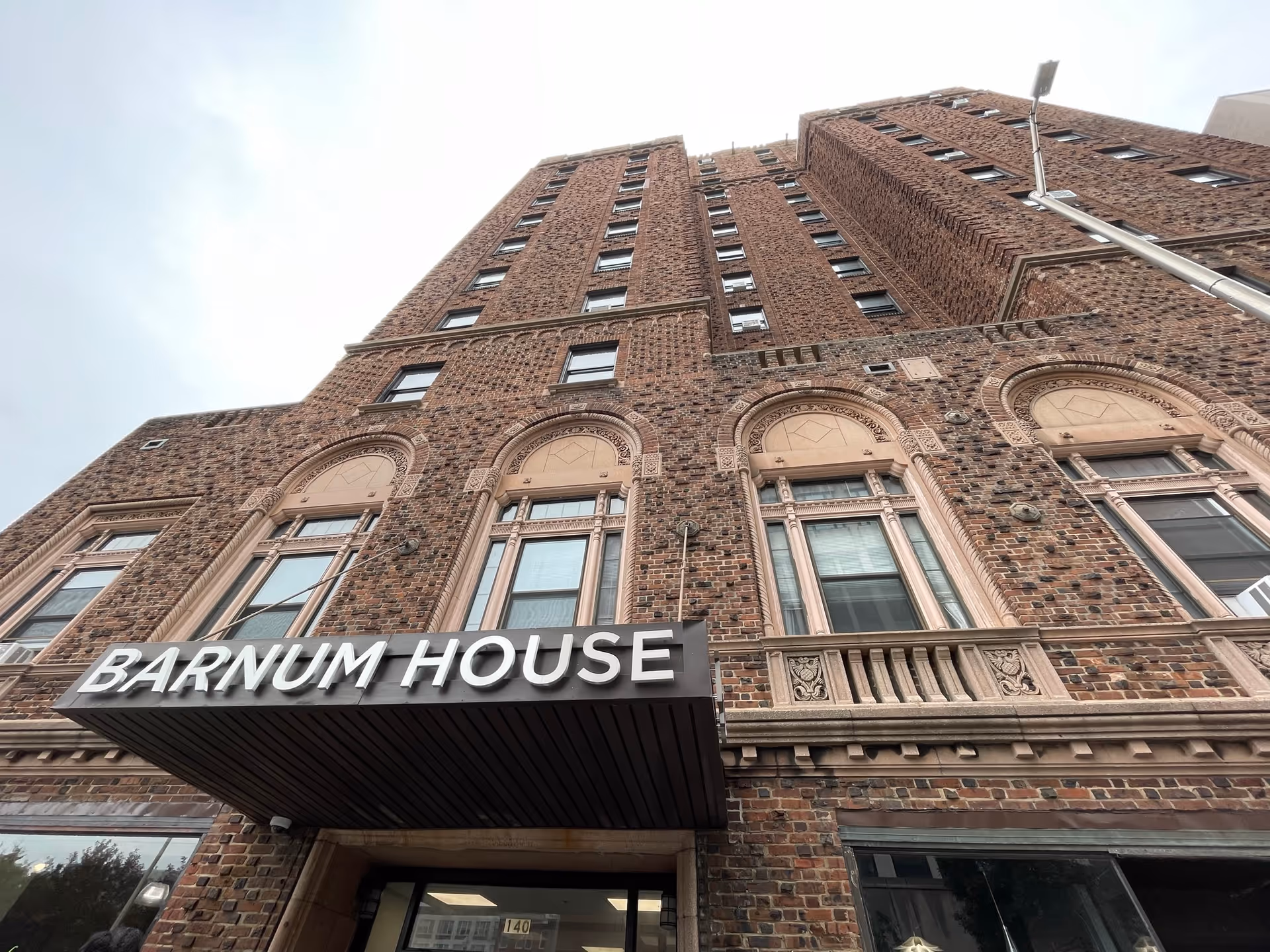 Upward view of a tall brick building with decorative stone window frames and an awning displaying the name 'Barnum House' above the entrance.