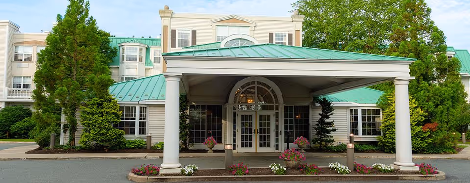 Front exterior view of a senior living facility with a green metal roof, white columns supporting a covered entrance, large windows, and landscaping with trees and flowers.