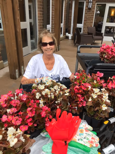 An elderly woman wearing sunglasses and a white Notre Dame t-shirt is smiling while sitting outdoors near a table filled with vibrant pink and white flowers and gardening gloves. The background shows a brick building with large windows and outdoor chairs.