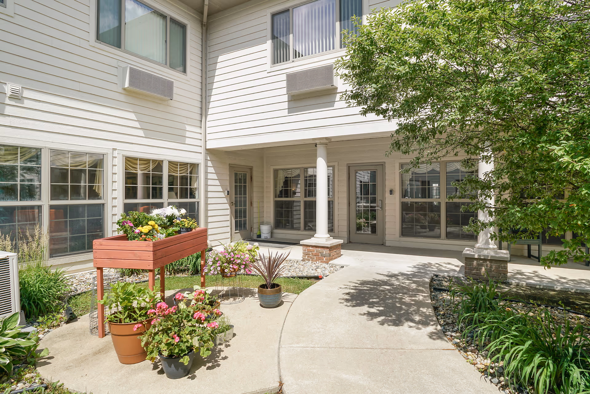 Outdoor courtyard area of a senior living facility with a concrete walkway, potted plants, a raised wooden planter box with flowers, and a building with large windows and doors in the background. There is greenery and a tree providing shade on the right side.