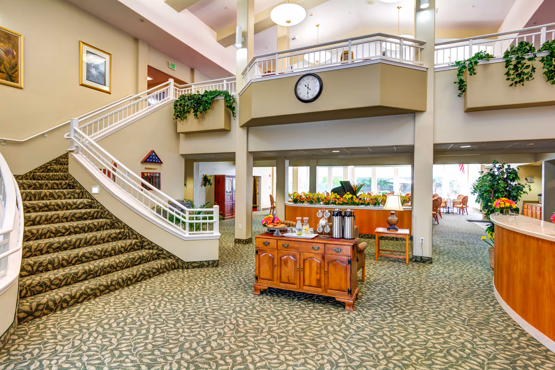 Bright senior living facility lobby with a carpeted grand staircase, central coffee/refreshment station, and seating areas under a mezzanine.