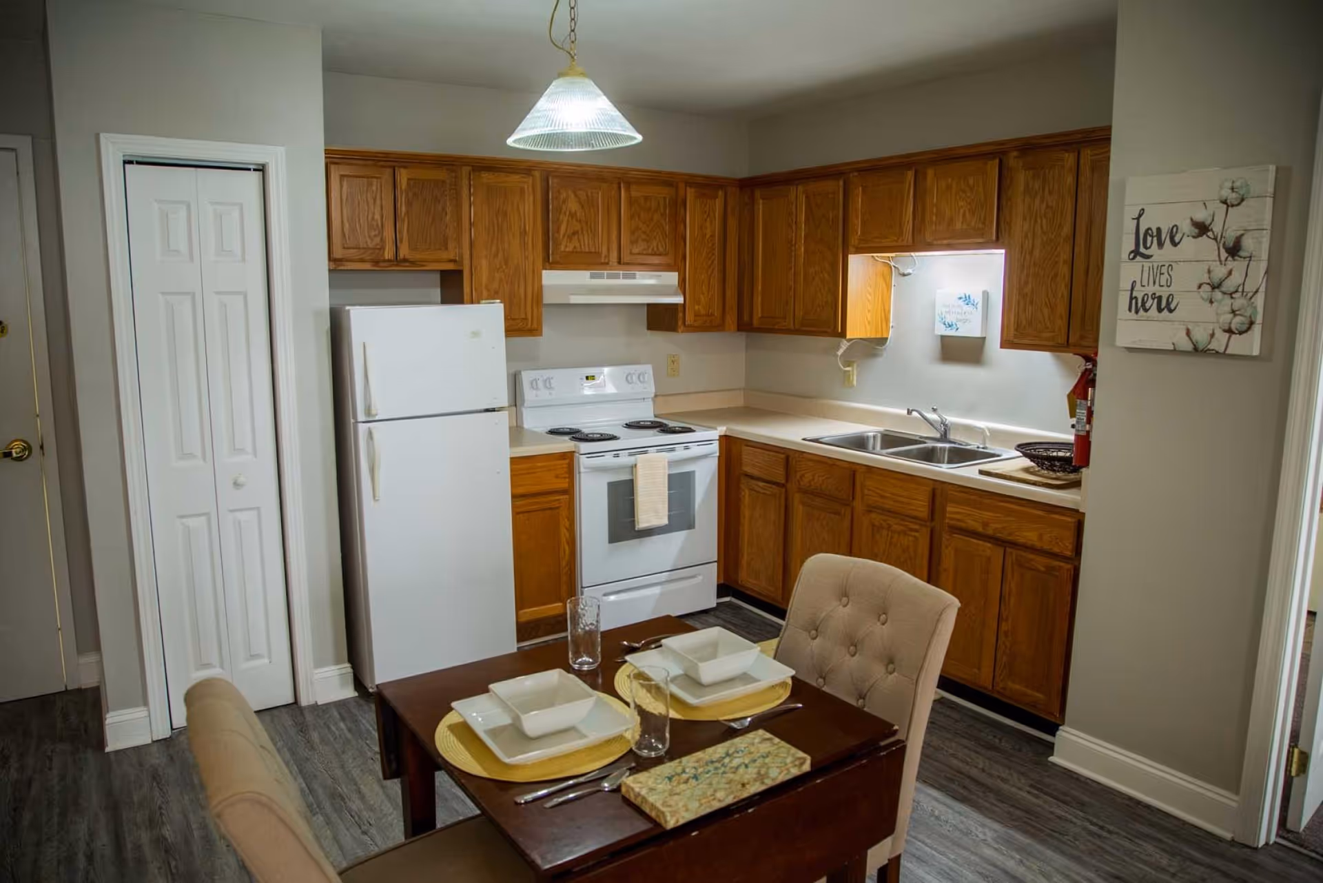 A kitchen with wooden cabinets, a white refrigerator, a white electric stove with an oven, and a double sink. A small dining table set for two with beige cushioned chairs is in the foreground. The walls are light gray, and there is a hanging light fixture above the table. A decorative sign on the wall reads 'Love lives here.'