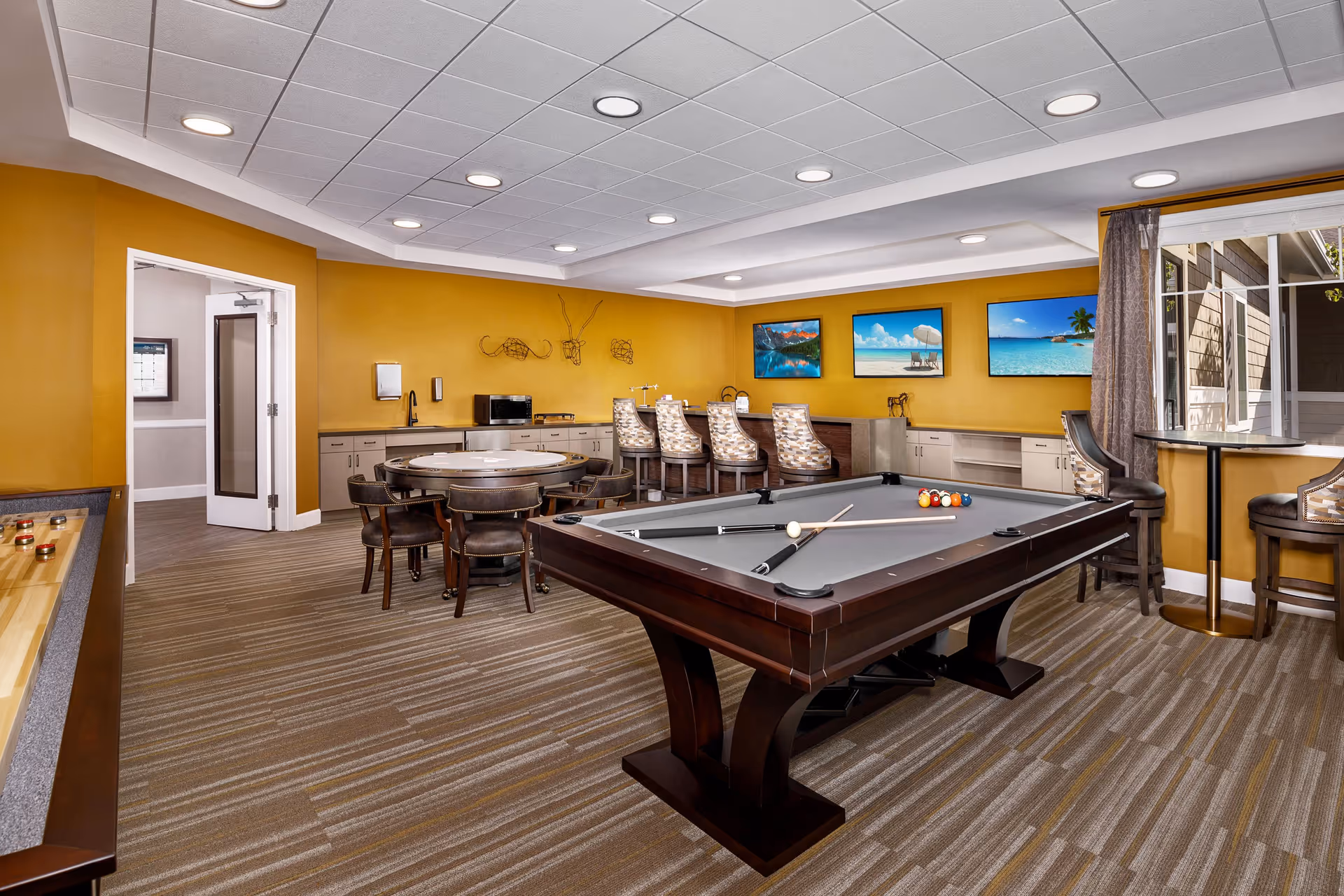 A recreational room with a pool table in the center, a round table with chairs, a shuffleboard table, and a bar counter with high chairs. The walls are painted mustard yellow and decorated with three framed pictures of beach and mountain scenes. There is a window with curtains on the right side letting in natural light.