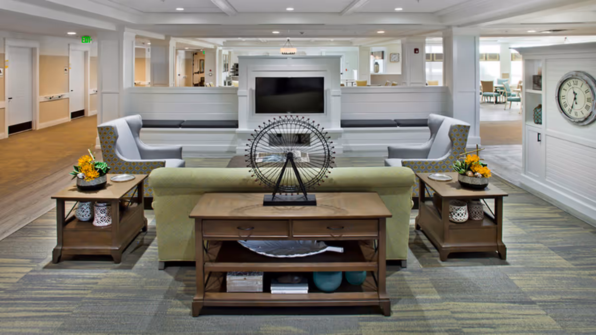 A cozy living room area in a senior living facility with a green sofa, two patterned armchairs, wooden side tables with flower arrangements, a flat-screen TV mounted on a white paneled wall, and a large clock on the right wall. The space is well-lit with natural light coming from windows in the background.