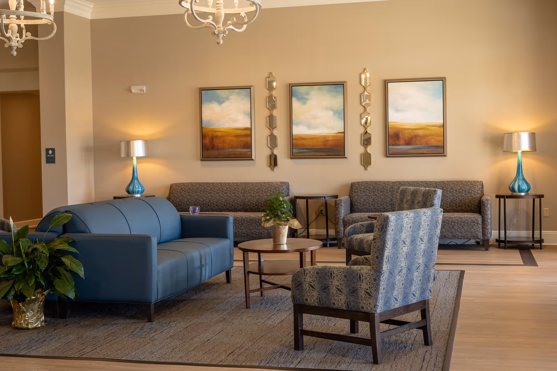 A cozy living room area with a blue leather sofa, two patterned armchairs, and two gray sofas. There is a round wooden coffee table with a potted plant in the center. The walls are beige with three landscape paintings and decorative mirrors hanging above the sofas. Two blue glass table lamps with silver shades are placed on side tables, and a green potted plant is on the floor near the sofa. The room has wooden flooring and two chandeliers hanging from the ceiling.