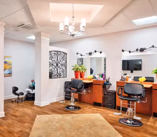Interior view of a salon area with two black salon chairs in front of mirrors mounted on a wooden counter. The room has wooden flooring, white walls, a ceiling light fixture, and decorative elements including potted plants and a wrought iron wall decoration.