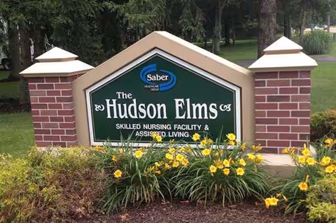Exterior sign for The Hudson Elms nursing facility, framed by brick pillars and yellow flowers.
