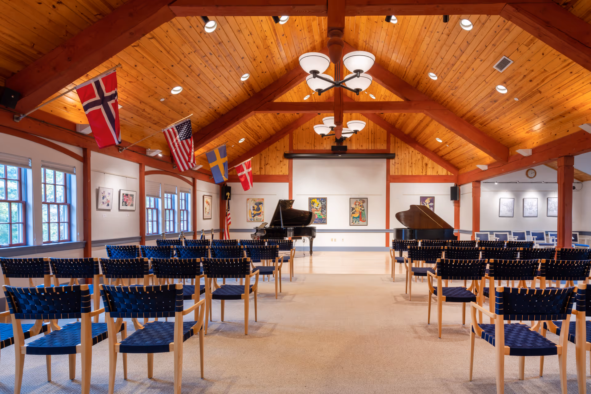 Interior view of a wooden-beamed assembly room with rows of chairs facing two grand pianos and a projection screen, decorated with hanging flags.