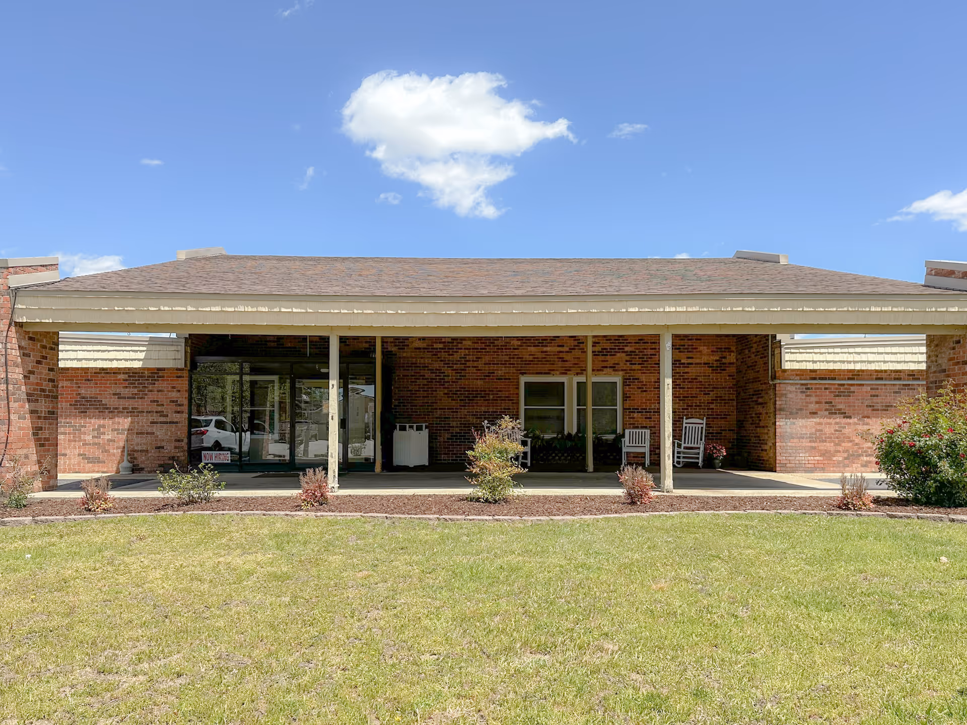 Covered brick front entrance of a single-story senior living facility with glass doors, porch rocking chairs and a lawn in front.