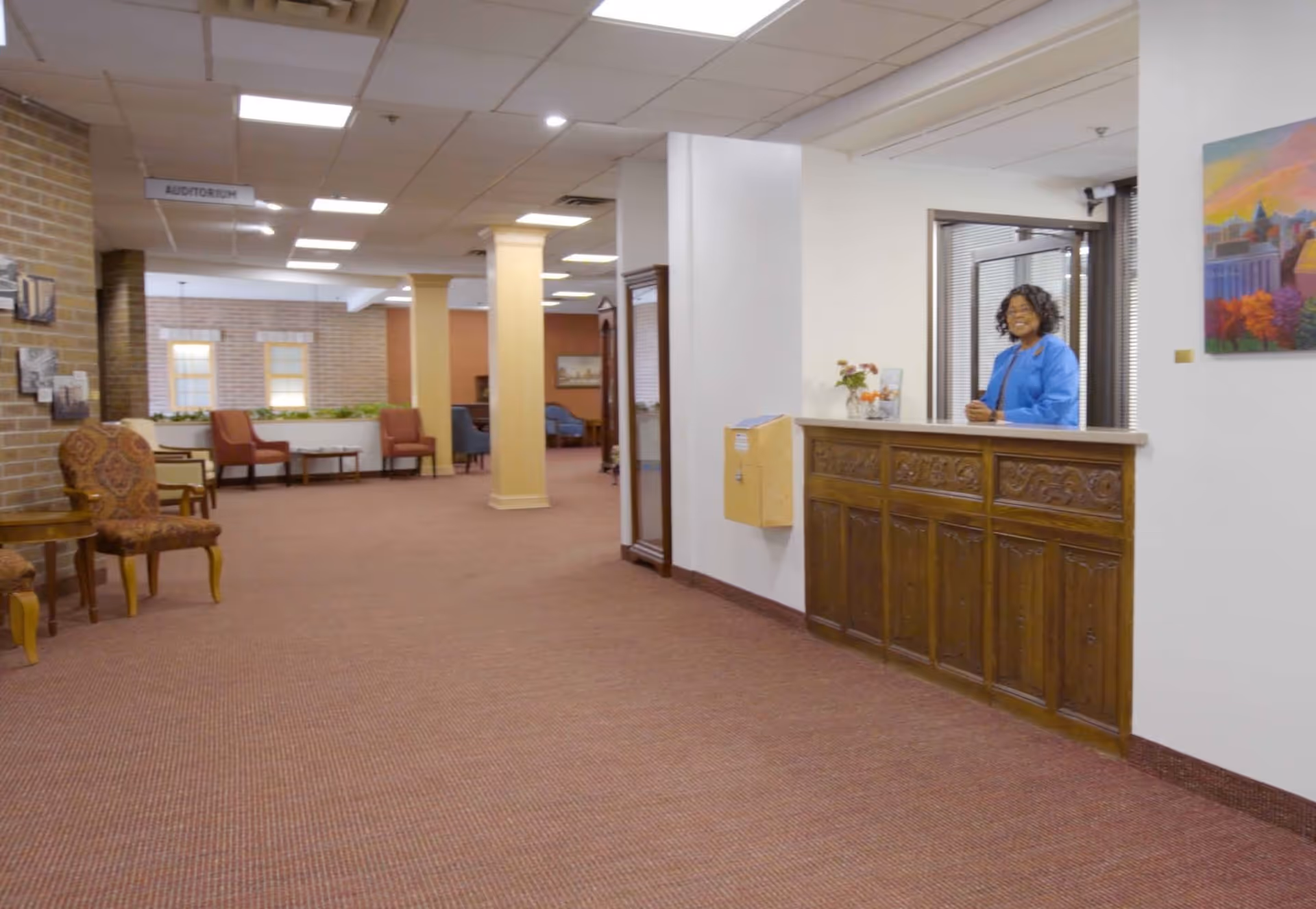 Interior view of a senior living facility hallway with carpeted floor, brick and white walls, and ceiling lights. There is a wooden reception desk on the right with a smiling woman standing behind it. Several chairs and small tables are arranged along the left side of the hallway. A sign hanging from the ceiling indicates the direction to the auditorium.