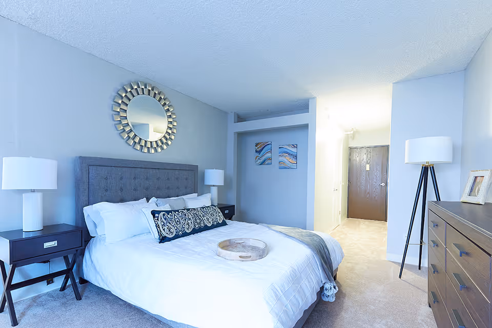 A bright and neatly arranged bedroom featuring a large bed with white bedding and decorative pillows. On either side of the bed are dark wooden nightstands with white lamps. Above the bed hangs a round decorative mirror. The room has light-colored walls and carpeted floor. To the right, there is a tall floor lamp next to a wooden dresser with a framed photo. In the background, a hallway leads to a closed wooden door, and two abstract paintings are mounted on the wall near the hallway.