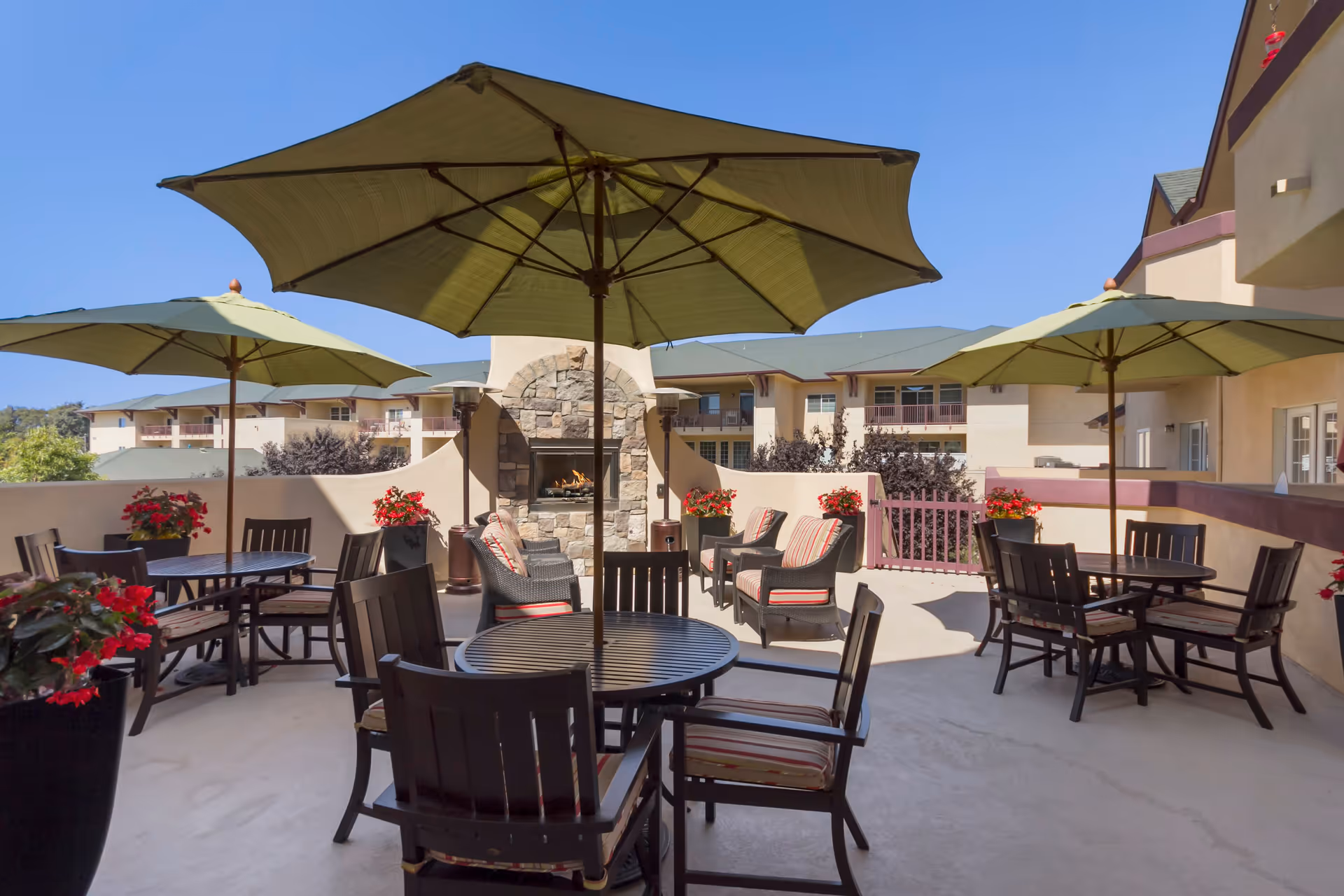 Outdoor patio area at Merrill Gardens At Santa Maria featuring several round tables with chairs and large green umbrellas. There are cushioned lounge chairs near a stone fireplace with a fire burning. Potted red flowers decorate the space, and the building exterior is visible in the background under a clear blue sky.