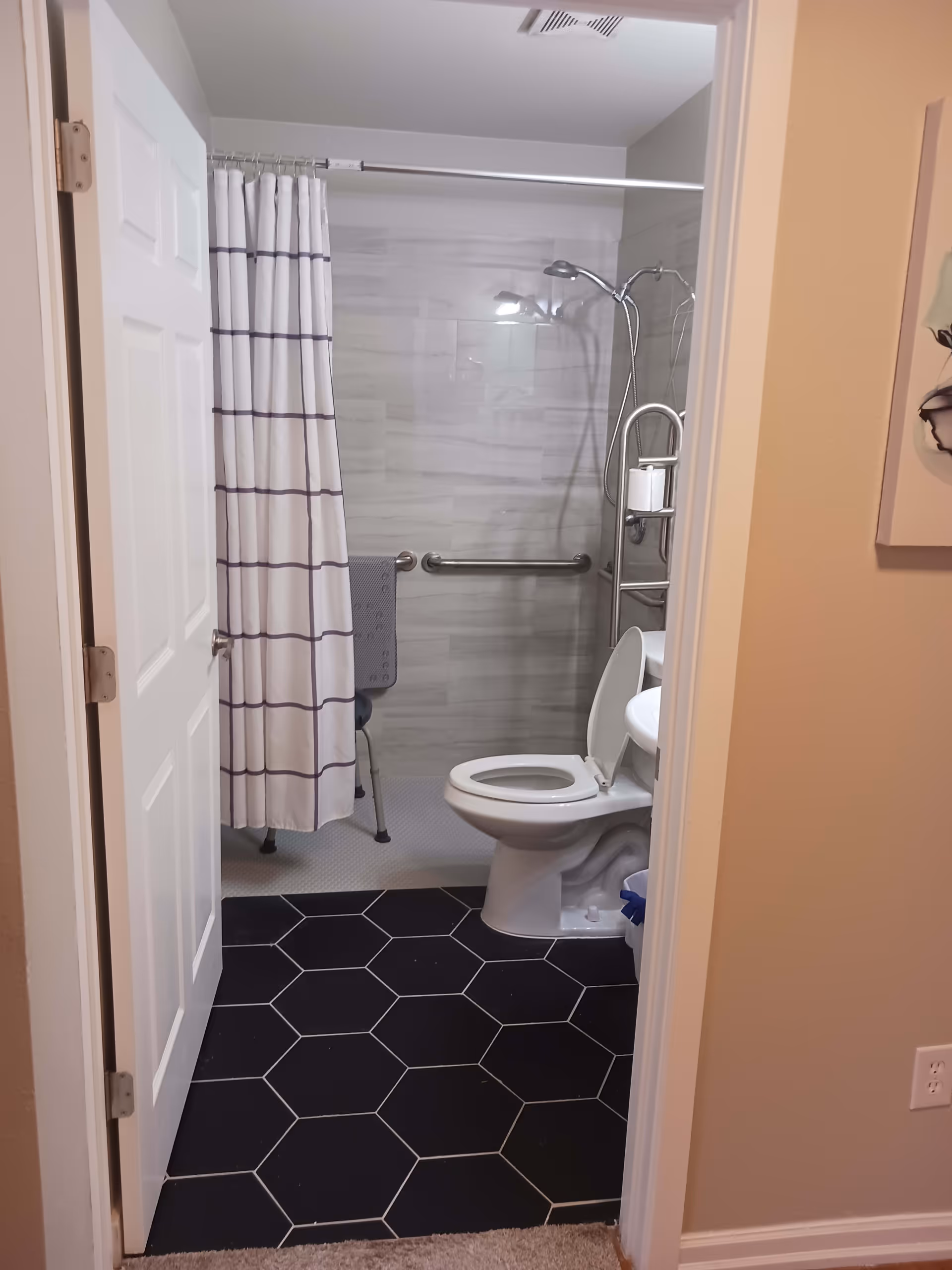 View into a bathroom with a white door open. Inside, there is a toilet with the lid up, a shower area with a white curtain featuring horizontal stripes, a shower chair, and a grab bar on the wall. The floor has black hexagonal tiles near the entrance and light-colored tiles in the shower area.