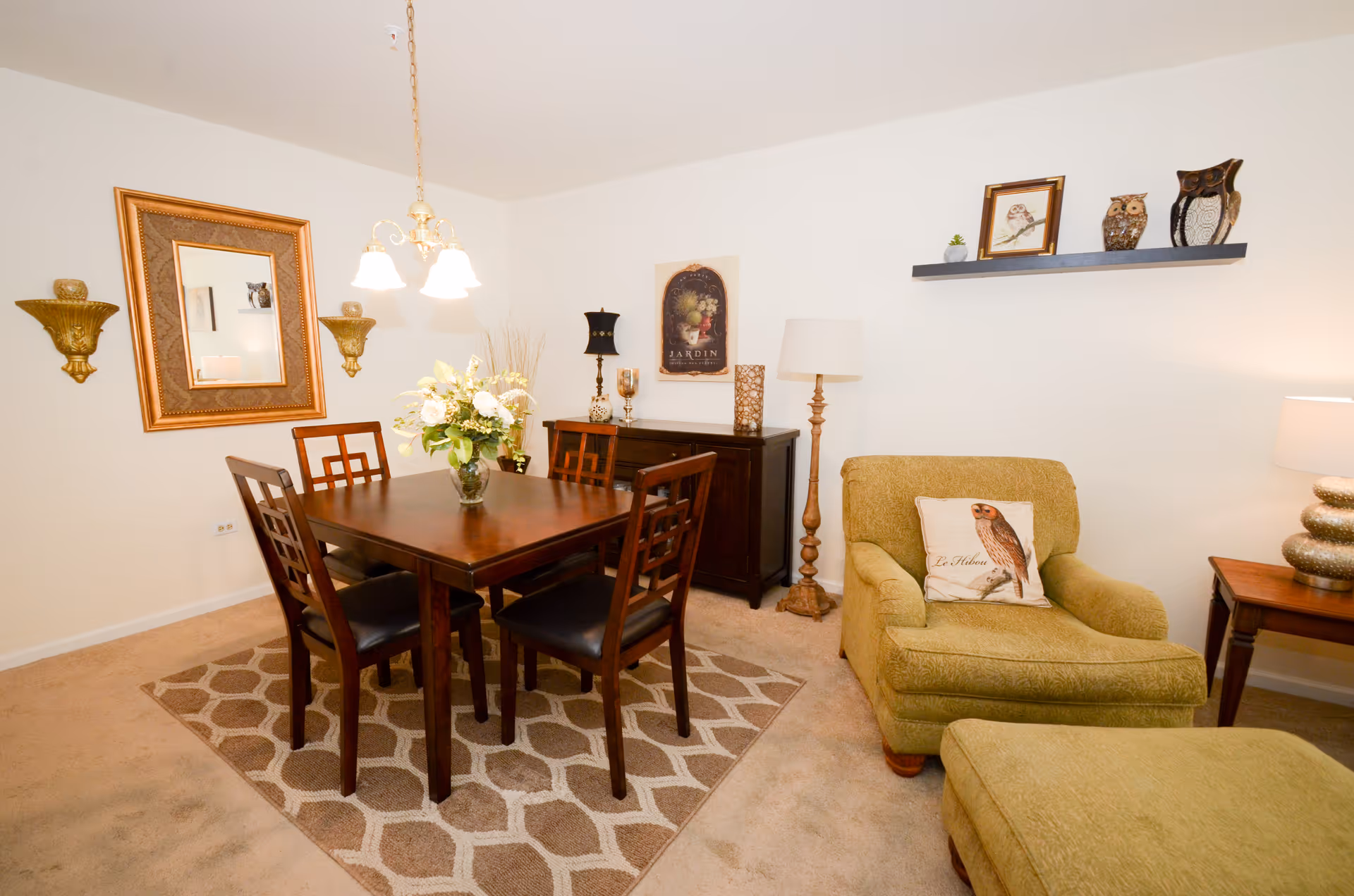 A cozy dining and sitting area featuring a wooden dining table with four chairs on a patterned rug, a green upholstered armchair with an owl-themed pillow, matching ottoman, a wooden sideboard with decorative items, a floor lamp, and wall decorations including a large mirror, sconces, framed artwork, and a floating shelf with owl figurines.
