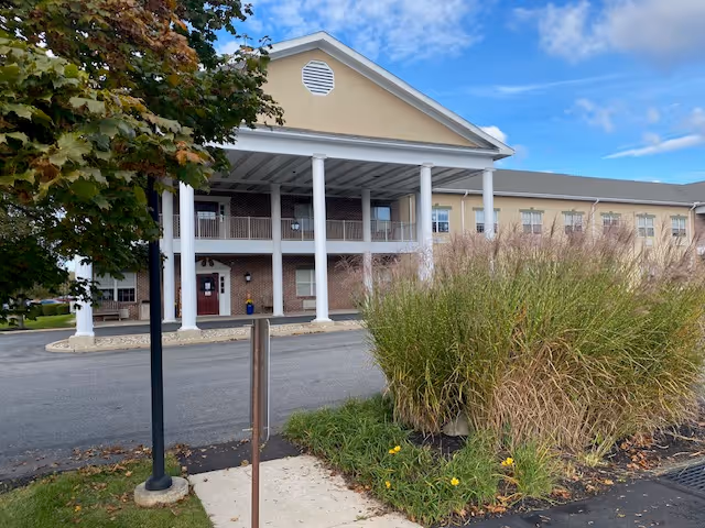 Front entrance of a beige senior living building with a covered portico supported by white columns, landscaped greenery, and a driveway.