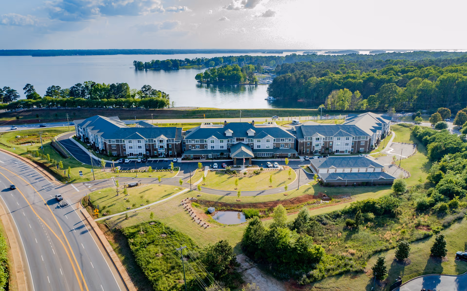 Aerial view of Lakeview Retirement Community, a large multi-story building complex surrounded by greenery and roads, with a lake and forested area in the background under a partly cloudy sky.
