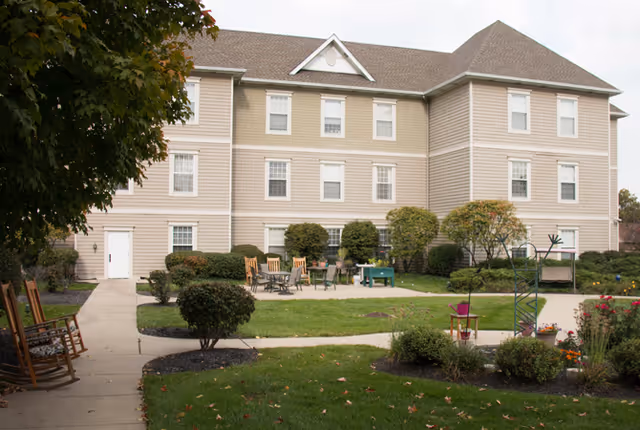 Outdoor garden area of a senior living facility with a beige three-story building in the background. The garden features a paved patio with several chairs and tables, surrounded by bushes, small trees, and well-maintained grass. There are rocking chairs along a sidewalk on the left side and various plants and flowers on the right.