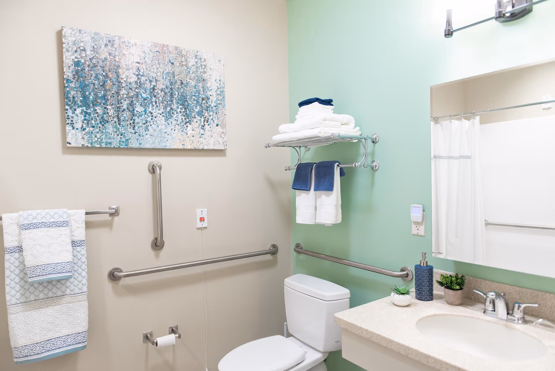 A clean and accessible bathroom with a white toilet, a countertop sink with a silver faucet, and a large mirror above. The walls are painted in beige and light green. There are multiple grab bars installed around the toilet area for support. A towel rack with folded white and blue towels is mounted on the wall, along with a shelf holding additional towels. A piece of abstract artwork with blue and white tones hangs on the beige wall.