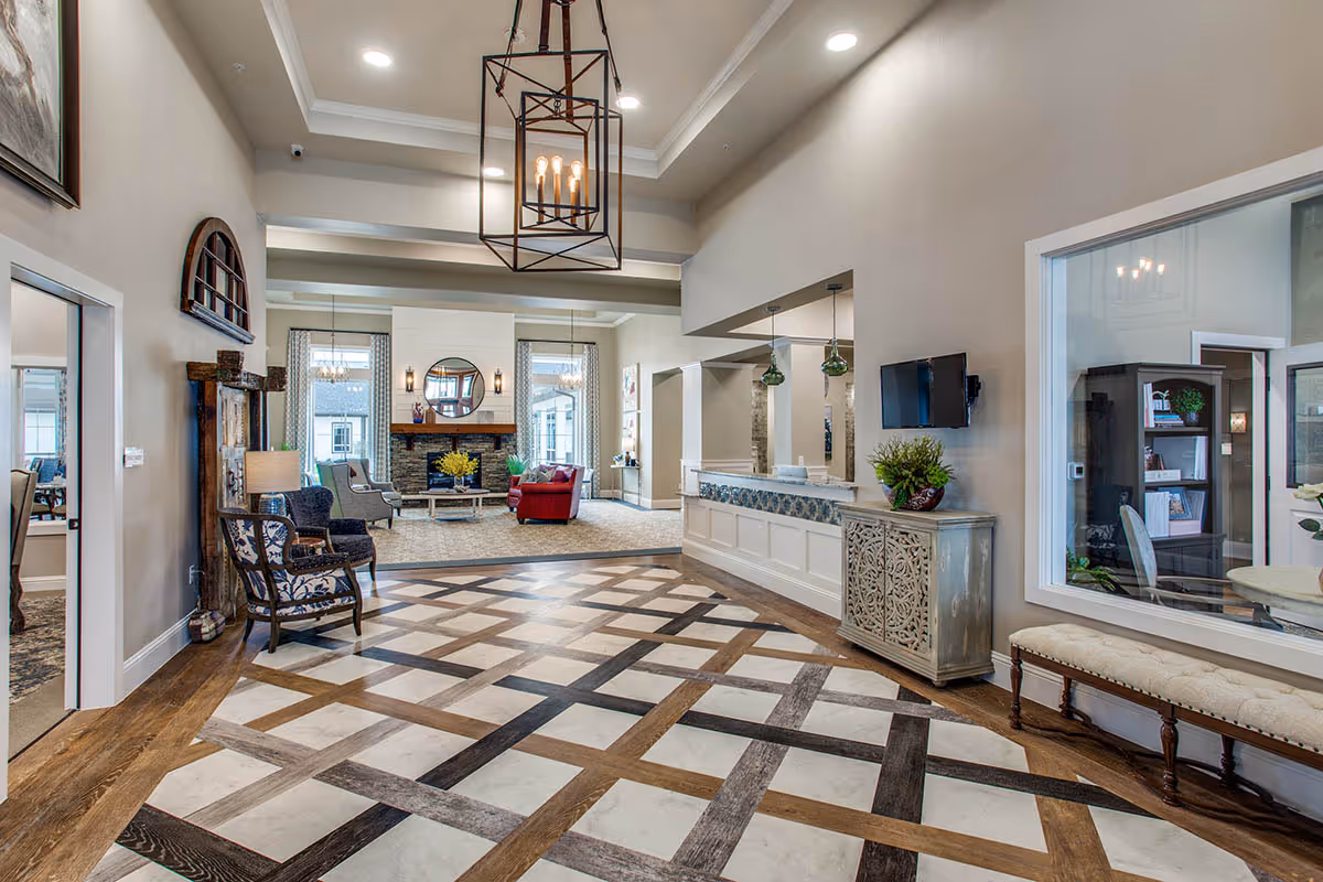 A spacious and well-lit senior living facility common area with a patterned floor, comfortable seating including armchairs and a red sofa, a fireplace with a round mirror above it, and a reception desk with decorative plants. The room features high ceilings with modern light fixtures and large windows with curtains.