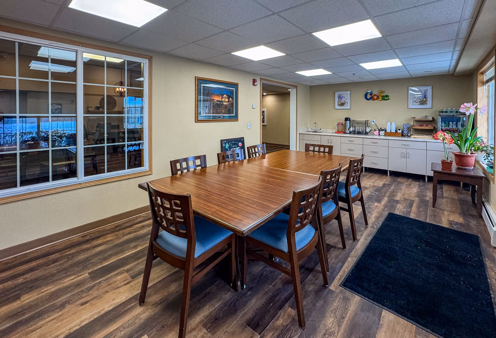 A dining area with a large wooden table surrounded by eight chairs with blue cushions. The room has wood flooring, beige walls, and a drop ceiling with fluorescent lights. On one side, there is a long counter with cabinets underneath, a coffee station, and decorative items including framed pictures and a colorful 'Cafe' sign on the wall. A window on the right side lets in natural light, and there are potted plants on a small table near the window. Another window on the left wall looks into an adjacent room.