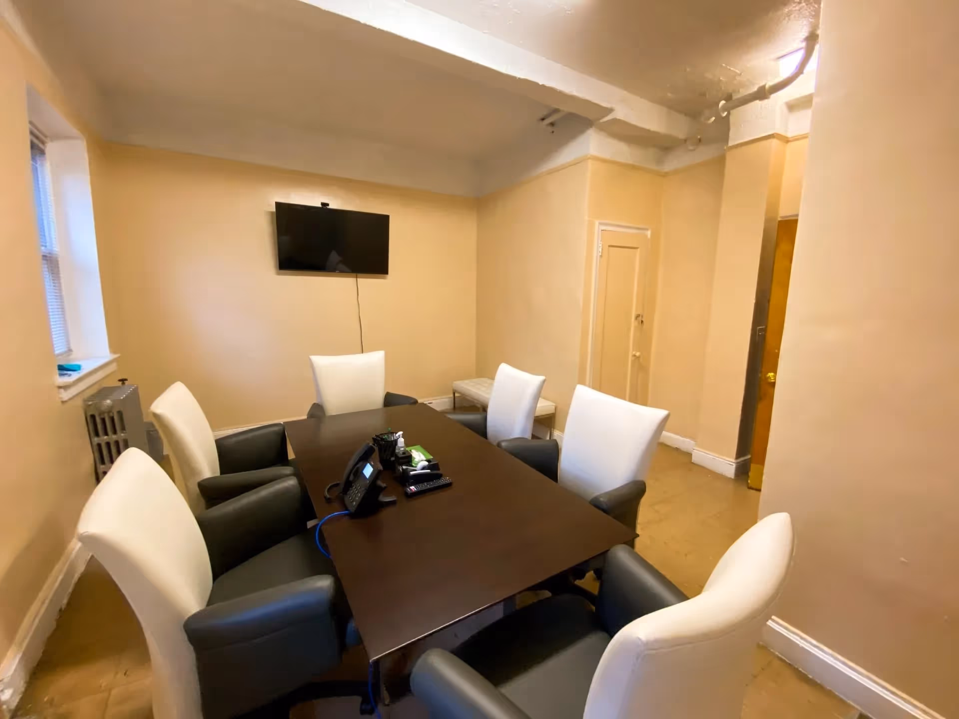 A small conference room with a dark wooden table surrounded by six chairs with white backs and black armrests. A wall-mounted flat screen TV is on the beige wall opposite a window with blinds. There is a telephone and some office supplies on the table. The room has beige walls and a light brown floor.