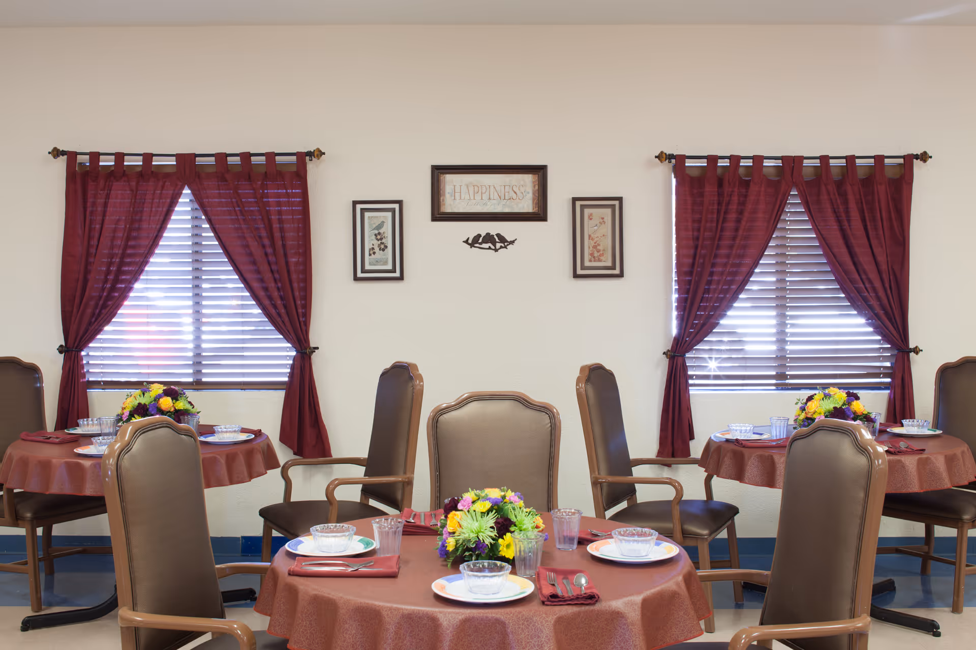 A dining room with round tables covered in red tablecloths, each set with plates, bowls, glasses, and silverware. Each table has a colorful flower centerpiece. The room has two windows with red curtains and blinds, and framed artwork on the wall between the windows.