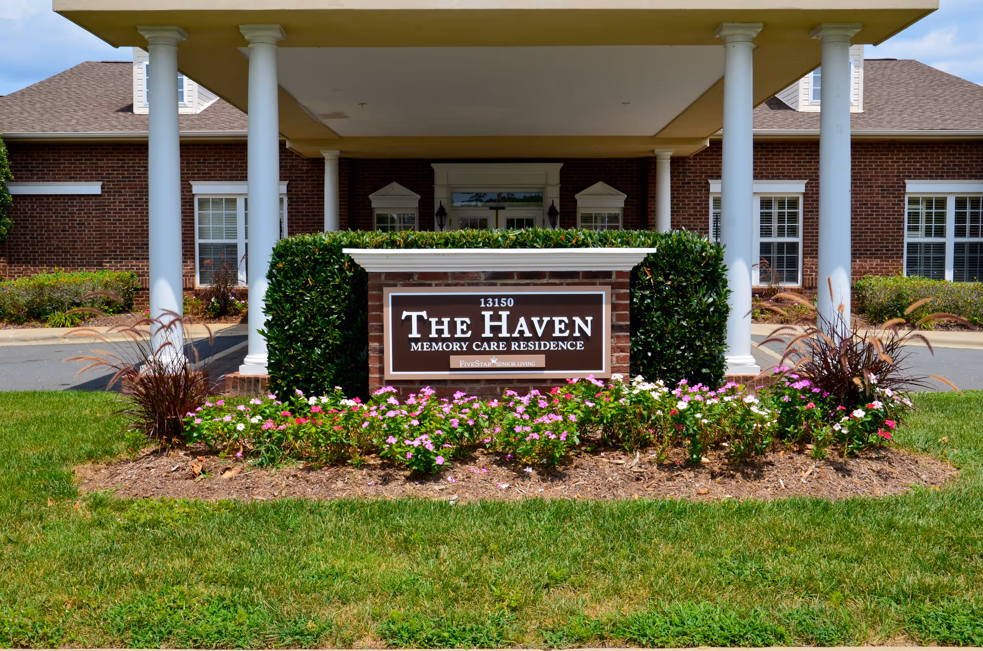Entrance of The Haven Memory Care Residence with a brick sign surrounded by flowers and greenery, supported by white columns and a building with windows in the background.