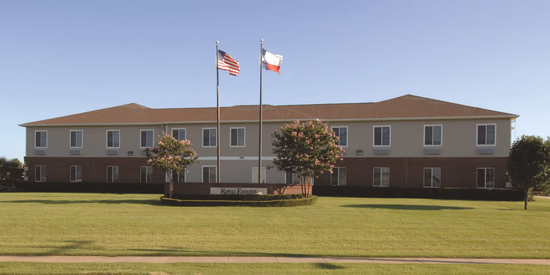 Exterior view of a two-story senior living facility building with a brown roof and beige and brick facade. Two flagpoles with the American flag and Texas state flag stand in front of the building, surrounded by neatly trimmed grass and small trees. A sign in front reads 'Royal Estates'.