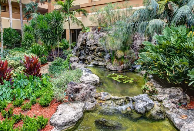 Lush indoor courtyard with a rock-lined pond and small waterfall surrounded by tropical plants and building balconies.