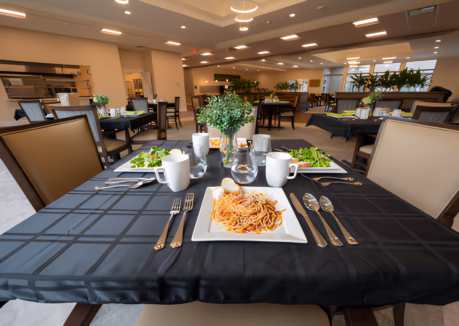 A dining table set for four with plates of food including spaghetti and salad, white mugs, glasses, and silverware on a black tablecloth. The background shows a spacious dining room with multiple tables and chairs, decorated with plants and modern lighting fixtures.