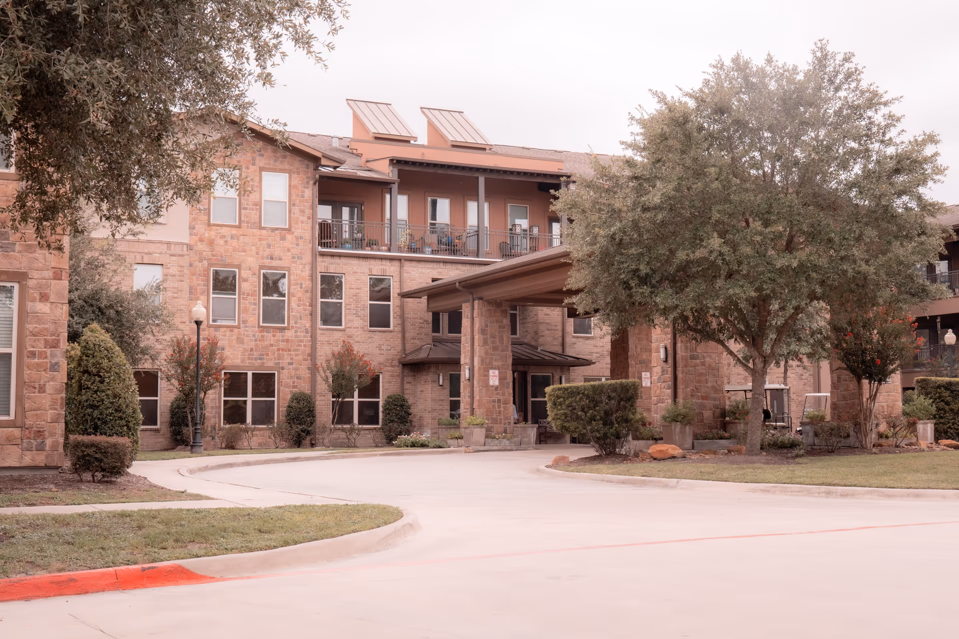 Front entrance of a multi-story brick building with a covered driveway, balconies, and landscaped trees.