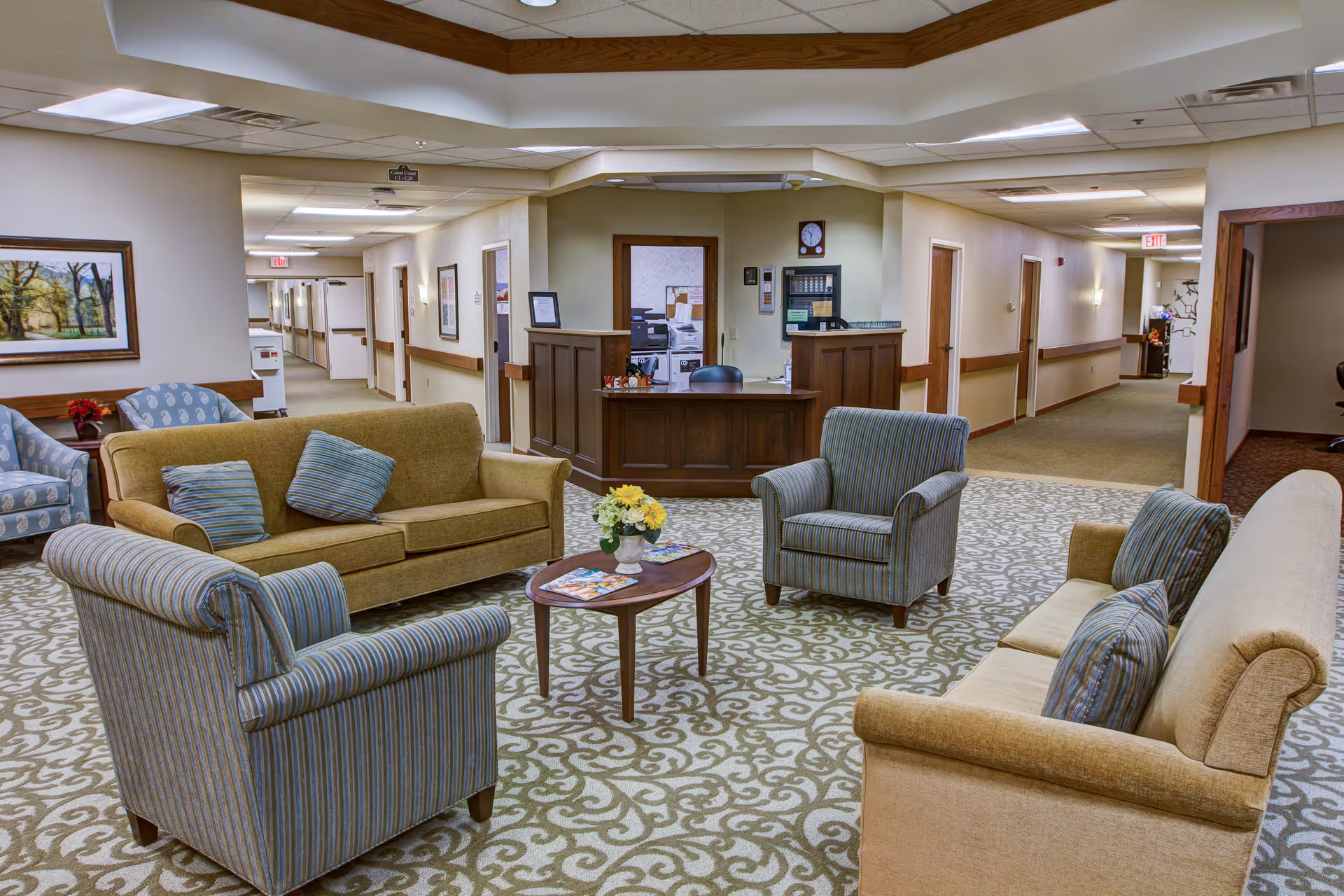 A furnished senior living lounge with sofas and armchairs arranged around a coffee table and a reception desk visible in the background.