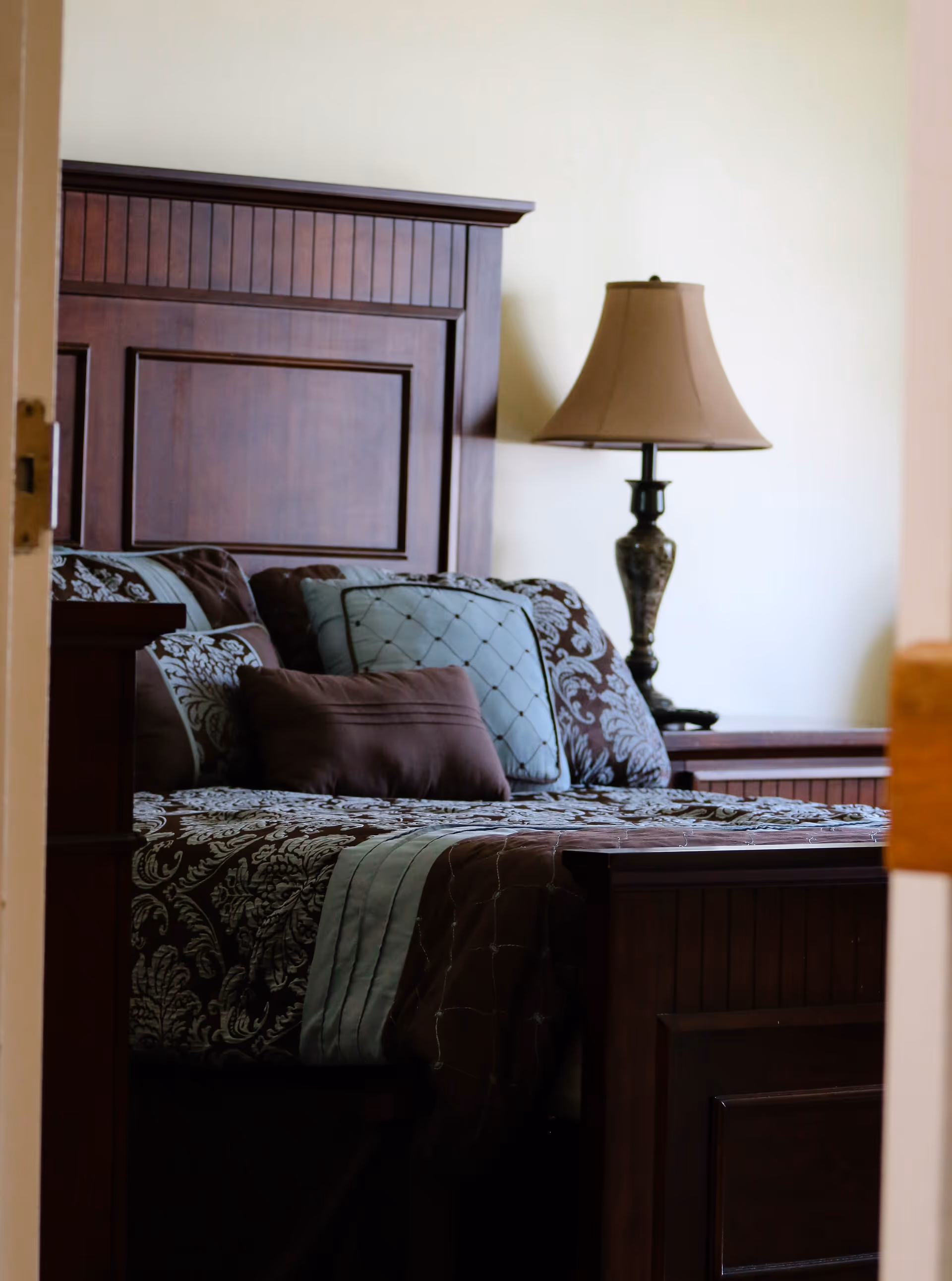 View through a partially open door into a bedroom featuring a wooden bed with an ornate headboard, decorated with patterned bedding and multiple pillows. A bedside table with a decorative lamp is visible next to the bed.
