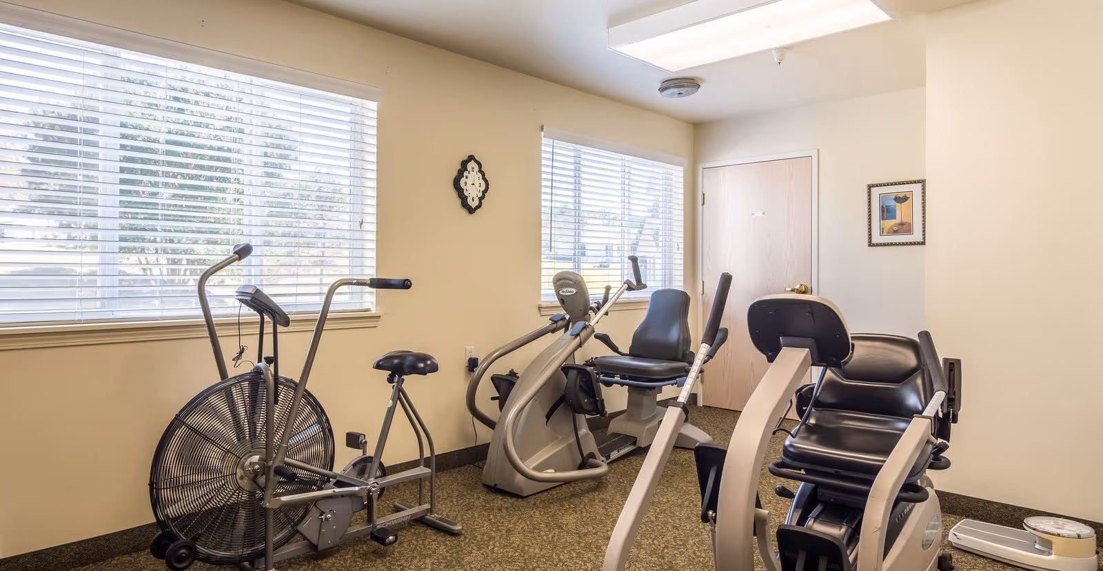Small fitness room with multiple exercise machines (recumbent bikes and an air bike) next to windows with blinds.