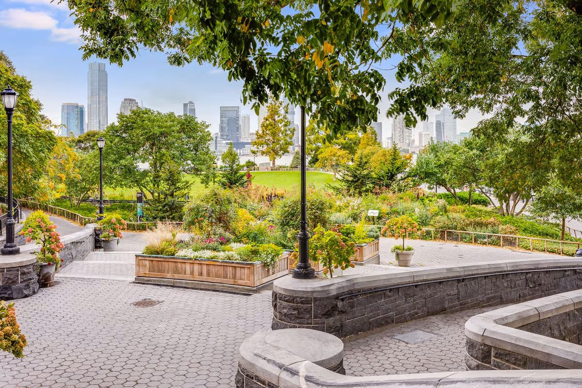 A landscaped outdoor garden area with paved walkways, stone walls, and various plants and trees. In the background, a city skyline with tall buildings is visible under a partly cloudy sky.