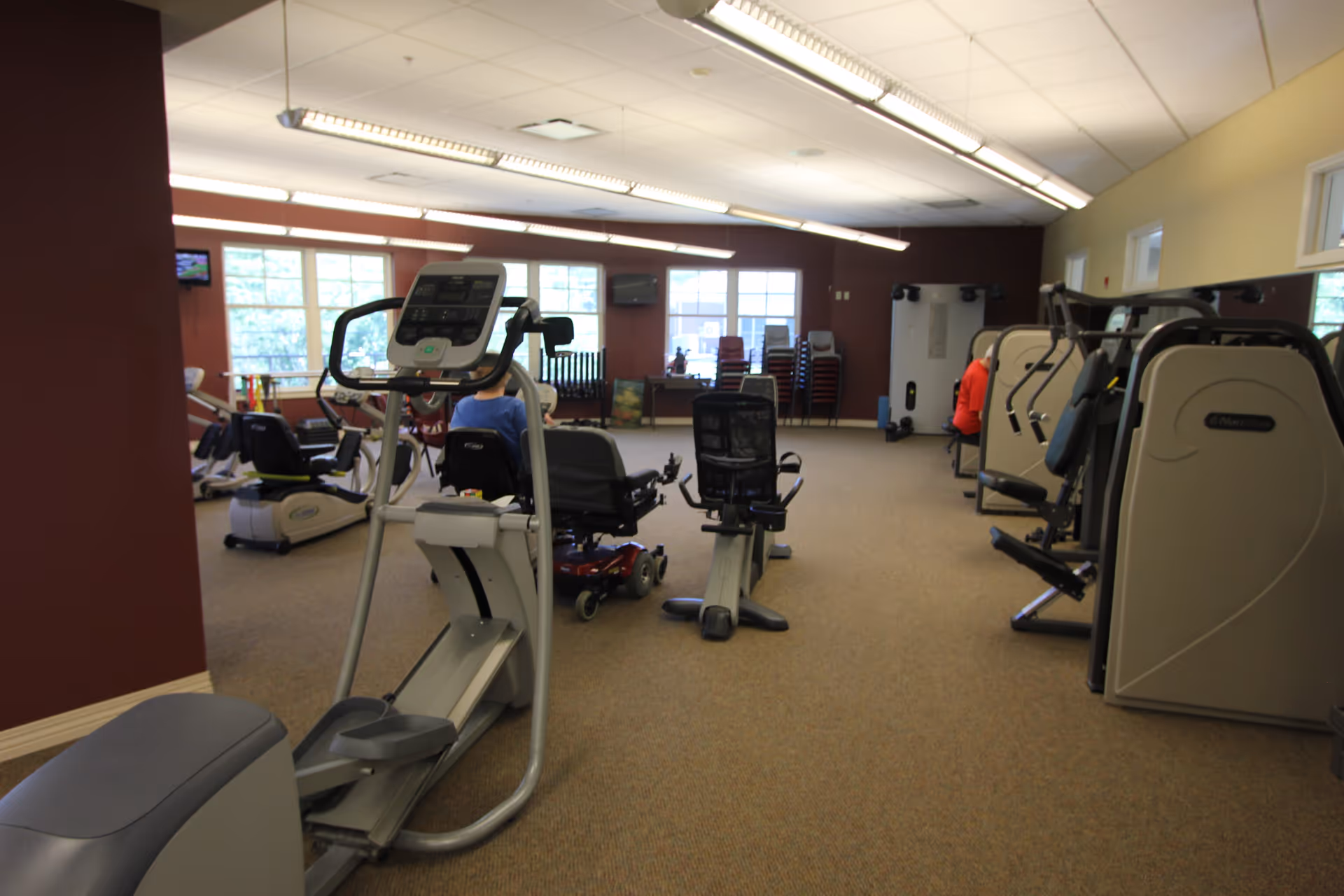 Exercise room with cardio and strength machines, chairs, and large windows.